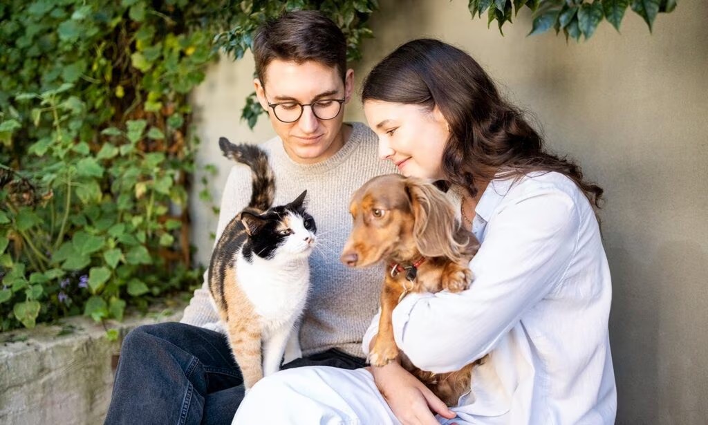 Young couple sitting outdoors with a calico cat and a dachshund, natural pet wellness UK